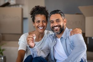 Woman with dark hair, wearing white polka dot shirt, smiling, and man, wearing blue shirt and white t-shirt, smiling, both facing forward, woman has her arm around man’s shoulder, man holding keys to their new home which they bought during the 2021 housing market