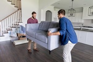 Two men, one with beard and wearing a red shirt, other wearing a blue short, former California residents move a sofa into their new home in Nevada